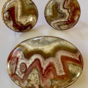 May include: A jewellery set comprising a brooch and two matching earrings. The brooch is oval, and the earrings are round. Each piece has a gold-coloured metal frame and a polished stone with red, white, and brown swirling patterns.