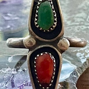 May include: A silver-toned ring with a vertical design, featuring a green stone at the top and a red stone at the bottom. The stones are set in black frames with a decorative edge. The ring has a horizontal bar and a rounded top and bottom.