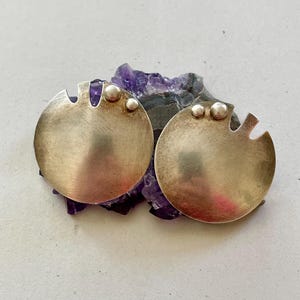 May include: A pair of circular silver earrings with a brushed metal finish, each featuring two small silver spheres and a notched edge detail, resting on a jagged piece of deep purple amethyst crystal with visible crystalline structures.