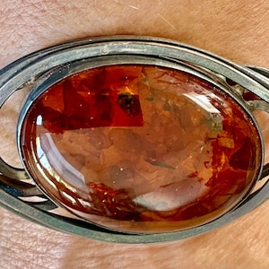 May include: Silver-tone cuff bracelet featuring a large, oval amber stone. The amber has a rich, reddish-brown color with internal inclusions. The bracelet has a decorative, openwork design.