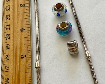 May include: A silver-toned necklace with a snake chain design, alongside two decorative glass beads and a small silver charm. The beads feature colorful patterns. A wooden ruler is placed next to the jewelry.