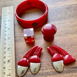 May include: A selection of vintage red accessories, including a bangle bracelet, a dice, a knob, and a pair of acorn-shaped earrings with silver-coloured bottoms. A wooden ruler is placed next to the items.