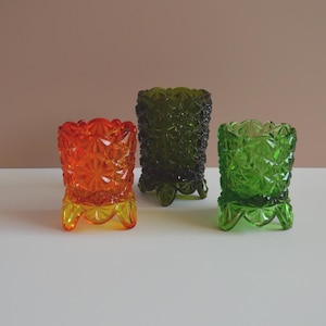 May include: Three vintage glass votive candle holders in orange, green, and dark green. Each has a textured, geometric pattern and sits on three small feet. The orange and green holders have a starburst design.