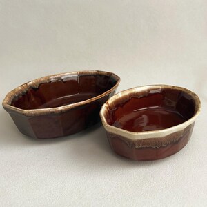 May include: Two oval-shaped ceramic bowls with a deep brown, glossy glaze and a lighter, cream-colored drip effect along the scalloped rims. The bowls feature a smooth, reflective interior surface and a sturdy, thick-walled construction.