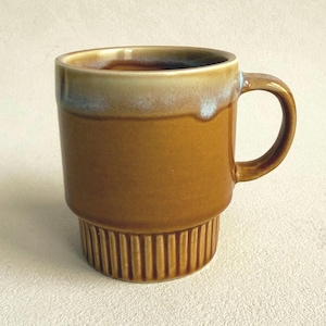 May include: A ceramic mug featuring a smooth, mustard-brown glaze on the upper body and a vertical ribbed texture on the lower base. The rim is finished with a lighter, creamy-white drip glaze effect, and the mug includes a curved side handle.