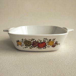 May include: A white ceramic baking dish with integrated side handles, featuring a decorative botanical print on the front. The design displays a colorful arrangement of various fruits and vegetables, including a red tomato, yellow lemon, and artichoke.