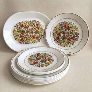 May include: A collection of white ceramic dinnerware featuring a central floral pattern in shades of orange, yellow, and green. The set includes one oval platter, one large round plate, and a stack of smaller round plates with brown rims.