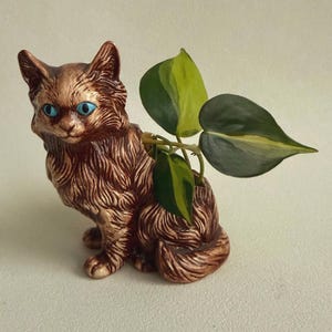 May include: A brown ceramic cat-shaped planter with textured fur details and bright blue eyes. The cat is in a seated position with a green and yellow variegated philodendron plant growing out of the opening on its back.