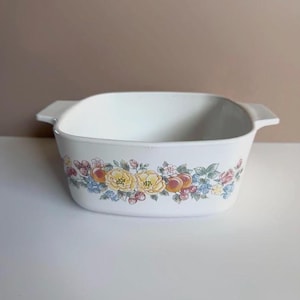 May include: White square baking dish with handles, decorated with a floral and fruit pattern. The design includes yellow flowers, red and orange fruits, and blue and pink accents. A vintage kitchenware item.