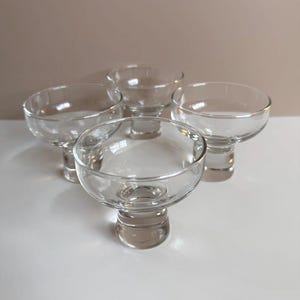 May include: Four clear glass coupe glasses with rounded bowls and short, pedestal stems. The glasses are arranged in a group, showcasing their simple, elegant design. The background is a neutral color, highlighting the glassware.
