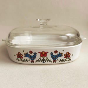 May include: A white rectangular casserole dish with a clear glass lid and a knob handle. The exterior features a colorful pattern of two blue birds facing a central red tulip, surrounded by green vines and small red and yellow flowers.