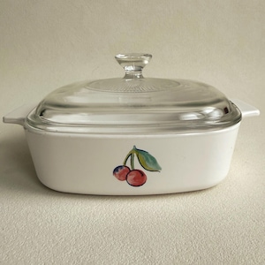 May include: A rectangular white casserole dish with integrated side handles and a clear glass lid featuring a central knob. The front of the dish is decorated with a painted illustration of two red cherries attached to a single green leaf stem.