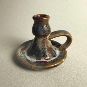 May include: A ceramic candle holder with a wide, circular base and a single curved handle. The piece features a mottled glaze in shades of brown, blue, white, and deep red, creating a variegated, glossy finish across the entire surface.