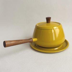 May include: A mustard yellow fondue pot with a matching lid and a long, straight wooden handle. The pot sits on a circular yellow base plate. The lid features a small, dark brown wooden knob at the center, and the handle is made of wood.