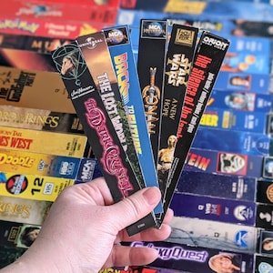 May include: A collection of VHS tapes with various movie titles, including "The Godfather", "Star Wars", "The Lost World", "Back to the Future", and "The Silence of the Lambs".