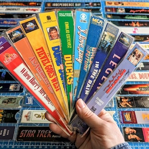 May include: A collection of VHS tapes fanned out in a hand. The tapes are mostly from the 1980s and 1990s and include titles like "Back to the Future", "When Harry Met Sally", "Dumbo", "Fievel Goes West", "An American Tail", "Ducks", "Legend", "In the Rain", and "Mrs. Doubtfire".