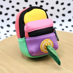 May include: A small, colorful backpack-shaped bag with a black zipper and a circular patch that reads "JOLLY HOUND". The bag is in shades of pink, yellow, purple, and green. A green bag is coming out of the bag.