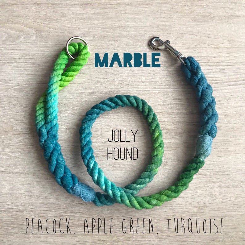 May include: A green, blue, and turquoise rope dog leash with a silver clip. The leash is tied in a circle and says "Jolly Hound" in the center. The text "MARBLE" is above the circle and the text "PEACOCK, APPLE GREEN, TURQUOISE" is below the circle.
