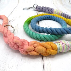 May include: A set of three colorful cotton rope dog leashes. The leashes are braided and feature a gradient of colors, including pink, orange, yellow, green, blue, and purple. The leashes are tied together with a pink and yellow striped ribbon.