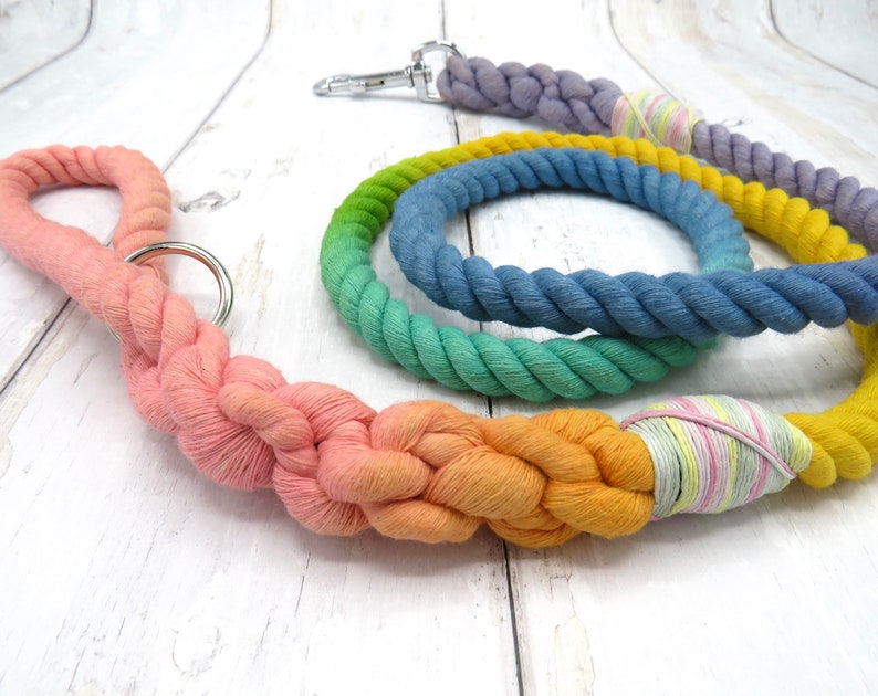 May include: A set of three colorful cotton rope dog leashes. The leashes are braided and feature a gradient of colors, including pink, orange, yellow, green, blue, and purple. The leashes are tied together with a pink and yellow striped ribbon.