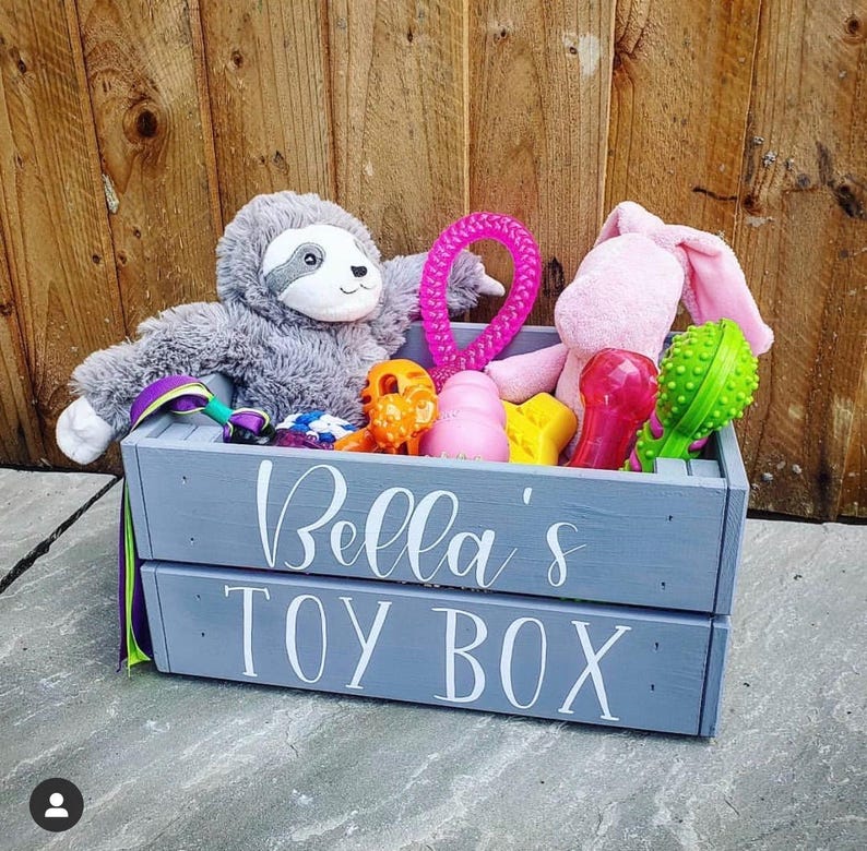 May include: A gray wooden toy box with the text "Bella's Toy Box" on the front. The box is filled with a variety of toys, including a pink plush bunny, a gray sloth, a pink and purple rope toy, and a variety of colorful chew toys.