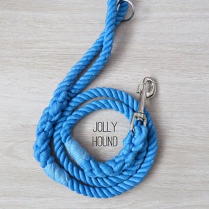 May include: A blue cotton rope dog leash with a silver clip and a ring at the top. The leash is coiled and the text "Jolly Hound" and "Seaside" are printed on the wooden surface.