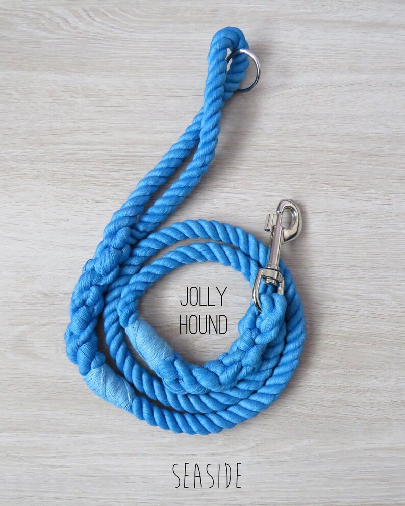 May include: A blue cotton rope dog leash with a silver clip and a ring at the top. The leash is coiled and the text "Jolly Hound" and "Seaside" are printed on the wooden surface.