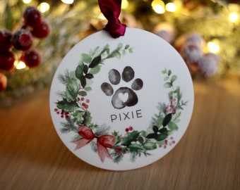 May include: Round white Christmas ornament with a green wreath, red bow, and a black paw print with a white heart in the center. The ornament is personalized with the name "PIXIE".