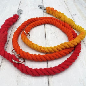 May include: Three dog leads in a gradient of colours, from red to orange to yellow. Each lead is made of thick, twisted rope with a metal clasp and a metal ring. The leads are arranged in a circular pattern on a light wood surface.
