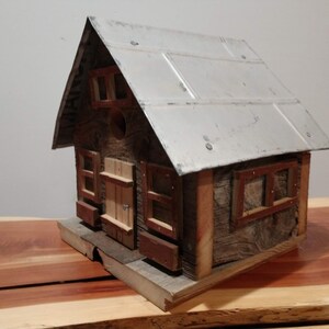 May include: A rustic wooden birdhouse with a metal roof. The birdhouse has a small door and windows with red trim. The wood has a weathered appearance, giving it a charming, country aesthetic. The birdhouse sits on a wooden base.