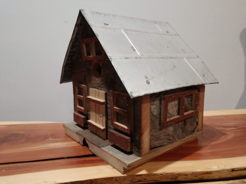 May include: A rustic wooden birdhouse with a metal roof. The birdhouse has a small door and windows with red trim. The wood has a weathered appearance, giving it a charming, country aesthetic. The birdhouse sits on a wooden base.