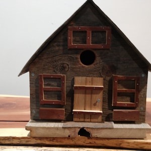 May include: A rustic wooden birdhouse with a gabled roof, a round entry hole, and small rectangular windows with red trim. The birdhouse is constructed from weathered wood, giving it a charming, aged appearance.