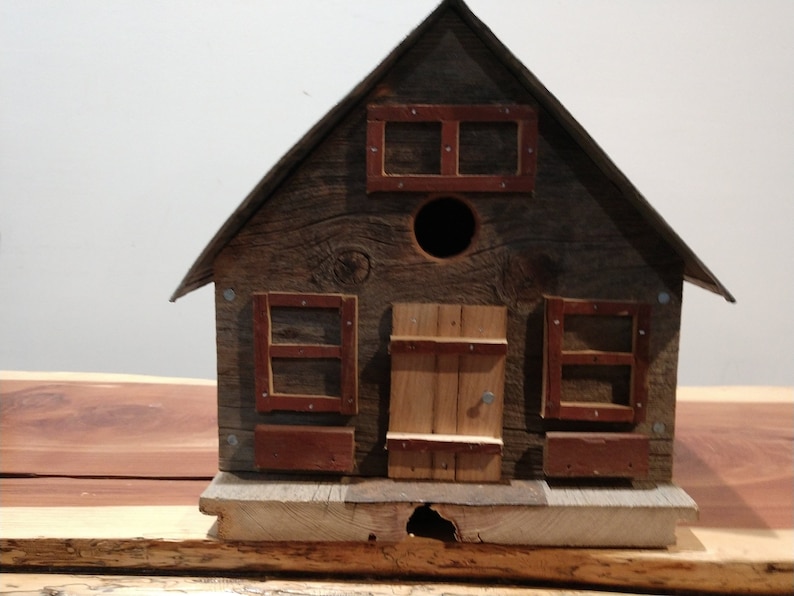 May include: A rustic wooden birdhouse with a gabled roof, a round entry hole, and small rectangular windows with red trim. The birdhouse is constructed from weathered wood, giving it a charming, aged appearance.