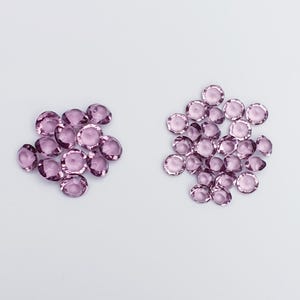 Puede incluir: Dos montones de pequeñas gemas de vidrio redondas y moradas sobre un fondo blanco.