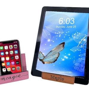 May include: Two phone stands, one pink and one brown, with personalized names "Meagen" and "Marle". The stands are made of leather and have a slot for holding a phone upright. The pink stand is holding a smartphone with a bright screen displaying a butterfly and the time 6:03.