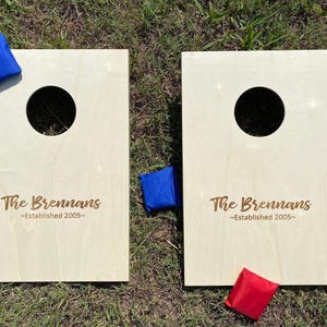 May include: Two wooden cornhole boards with a circular hole and the text "The Brennans ~Established 2005~". Includes blue and red bean bags. The boards are on a grassy surface.