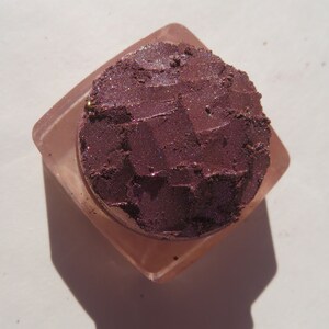 May include: A small, square, clear container with a lid, filled with a deep purple eyeshadow with a shimmery finish.