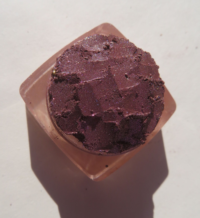 May include: A small, square, clear container with a lid, filled with a deep purple eyeshadow with a shimmery finish.