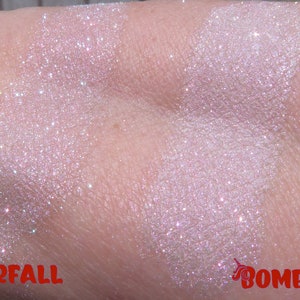 Op de afbeelding: Close-up van twee swatches van glinsterende, lichtroze oogschaduw op de huid. De linker swatch is gelabeld "Starfall" en de rechter swatch is gelabeld "Bombshell". Beide hebben een glitterende, iriserende finish.