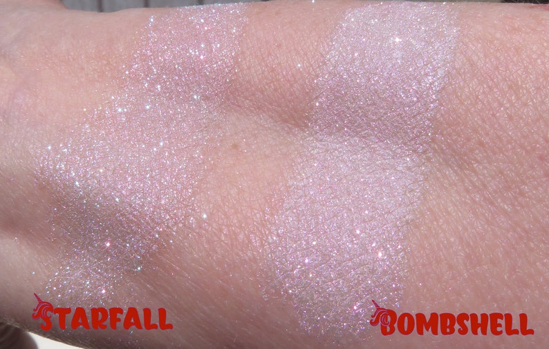 Op de afbeelding: Close-up van twee swatches van glinsterende, lichtroze oogschaduw op de huid. De linker swatch is gelabeld "Starfall" en de rechter swatch is gelabeld "Bombshell". Beide hebben een glitterende, iriserende finish.