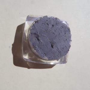 May include: A small, clear plastic container with a lid, filled with a dark gray eyeshadow powder.