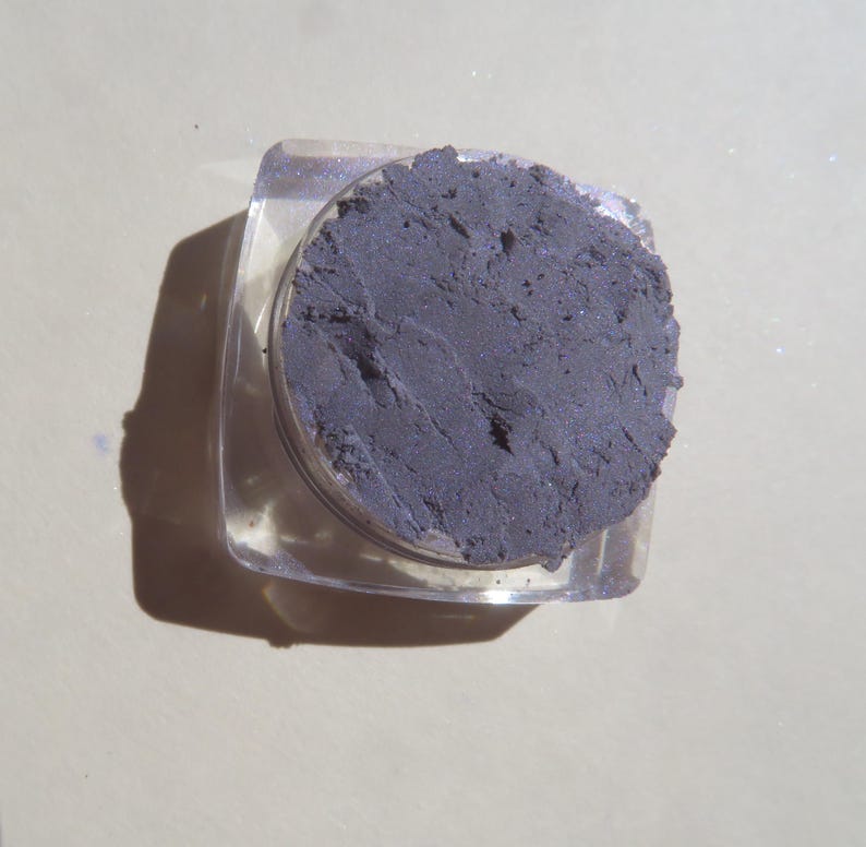 May include: A small, clear plastic container with a lid, filled with a dark gray eyeshadow powder.