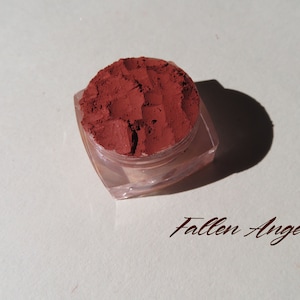 Op de afbeelding: Een kleine, heldere, vierkante container gevuld met een diep roodbruin poeder. De container staat op een wit oppervlak, met de woorden "Fallen Angel" in een cursief lettertype eronder. Het poeder lijkt een cosmetisch product te zijn.