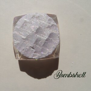 Op de afbeelding: Een vierkante, doorzichtige container gevuld met een glinsterend, licht zilverkleurig poeder. Het poeder heeft een textuur met een geometrisch patroon. Het woord "Bombshell" staat in cursief schrift onderaan.