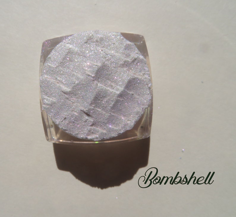 Op de afbeelding: Een vierkante, doorzichtige container gevuld met een glinsterend, licht zilverkleurig poeder. Het poeder heeft een textuur met een geometrisch patroon. Het woord "Bombshell" staat in cursief schrift onderaan.