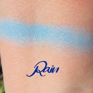May include: A light blue eyeshadow swatch on skin with the word "Rain" written in blue below it.