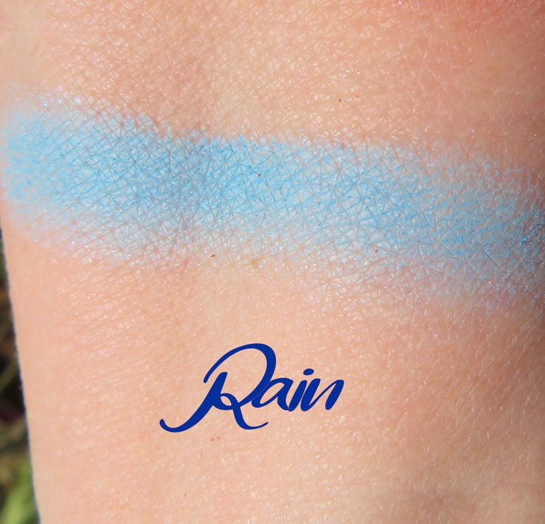 May include: A light blue eyeshadow swatch on skin with the word "Rain" written in blue below it.