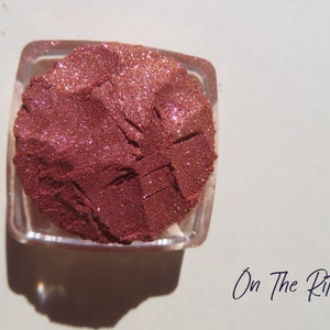 May include: A square, clear container filled with loose, shimmering copper-toned cosmetic powder. The surface of the powder shows distinct indentations and a fine, glittery texture. The text "On The Ritz" is printed in black near the container.