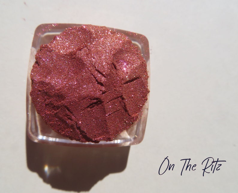 May include: A square, clear container filled with loose, shimmering copper-toned cosmetic powder. The surface of the powder shows distinct indentations and a fine, glittery texture. The text "On The Ritz" is printed in black near the container.