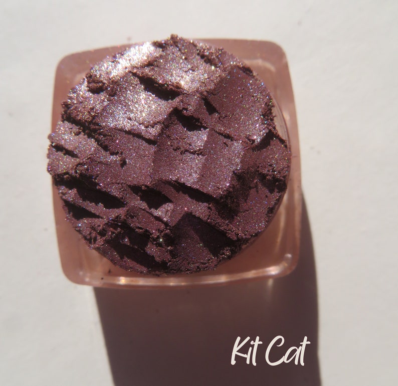 May include: A close-up of a small, round container of purple eyeshadow with a shimmery finish. The container is labeled "Kit Cat".